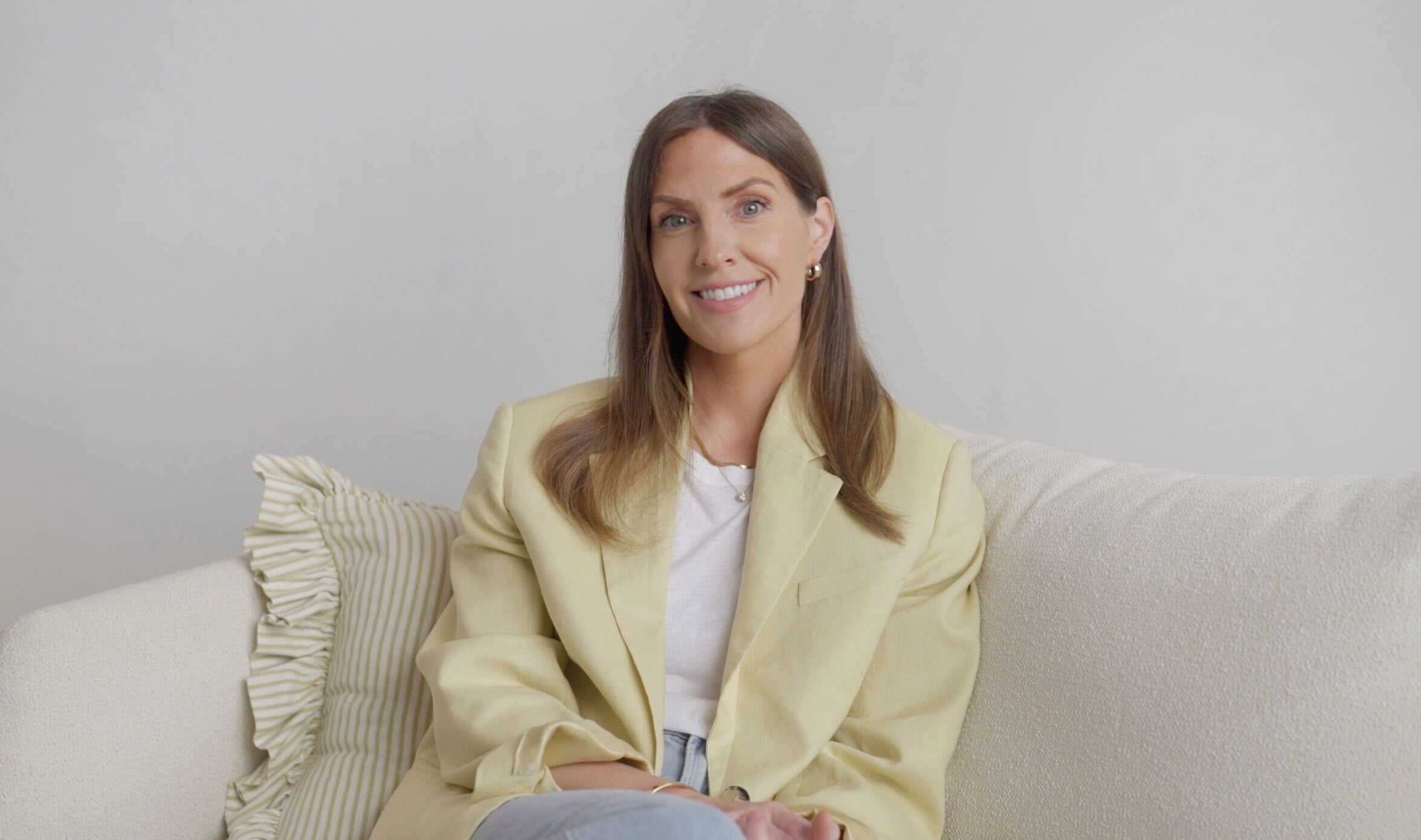 Woman sitting on a couch wearing a light-colored jacket against a plain background