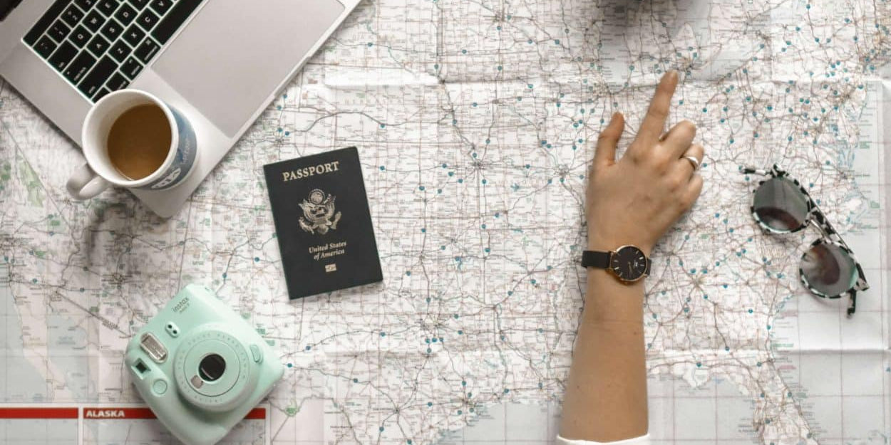 Laptop, passport, camera, and sunglasses on top of map with hand pointing at location