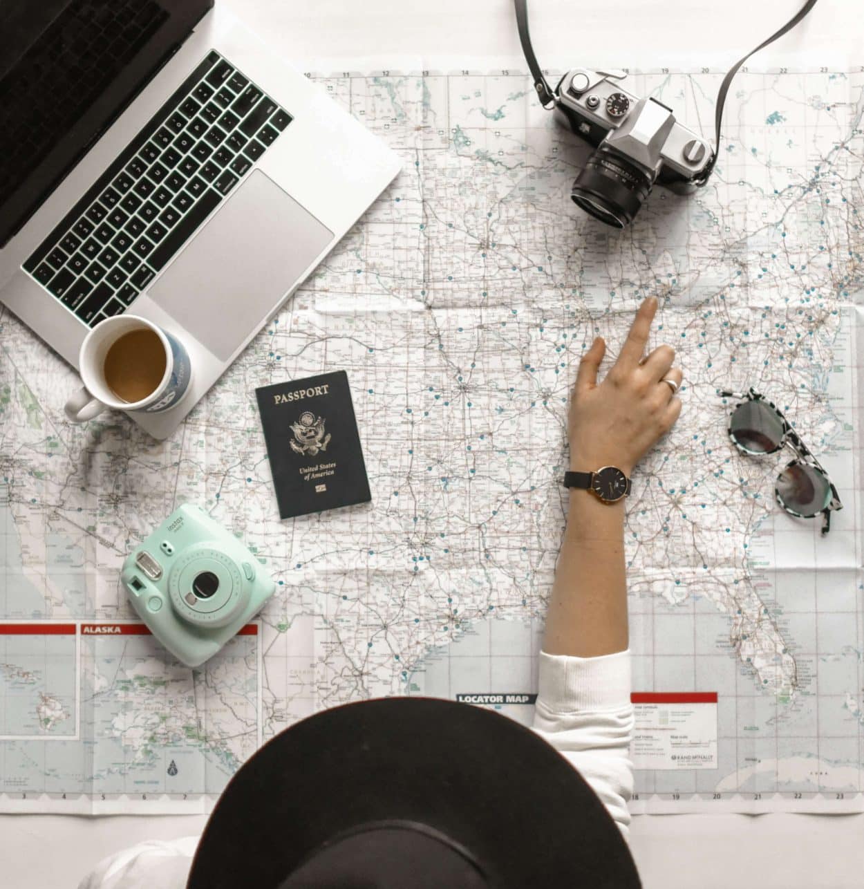 Laptop, passport, camera, and sunglasses on top of map with hand pointing at location