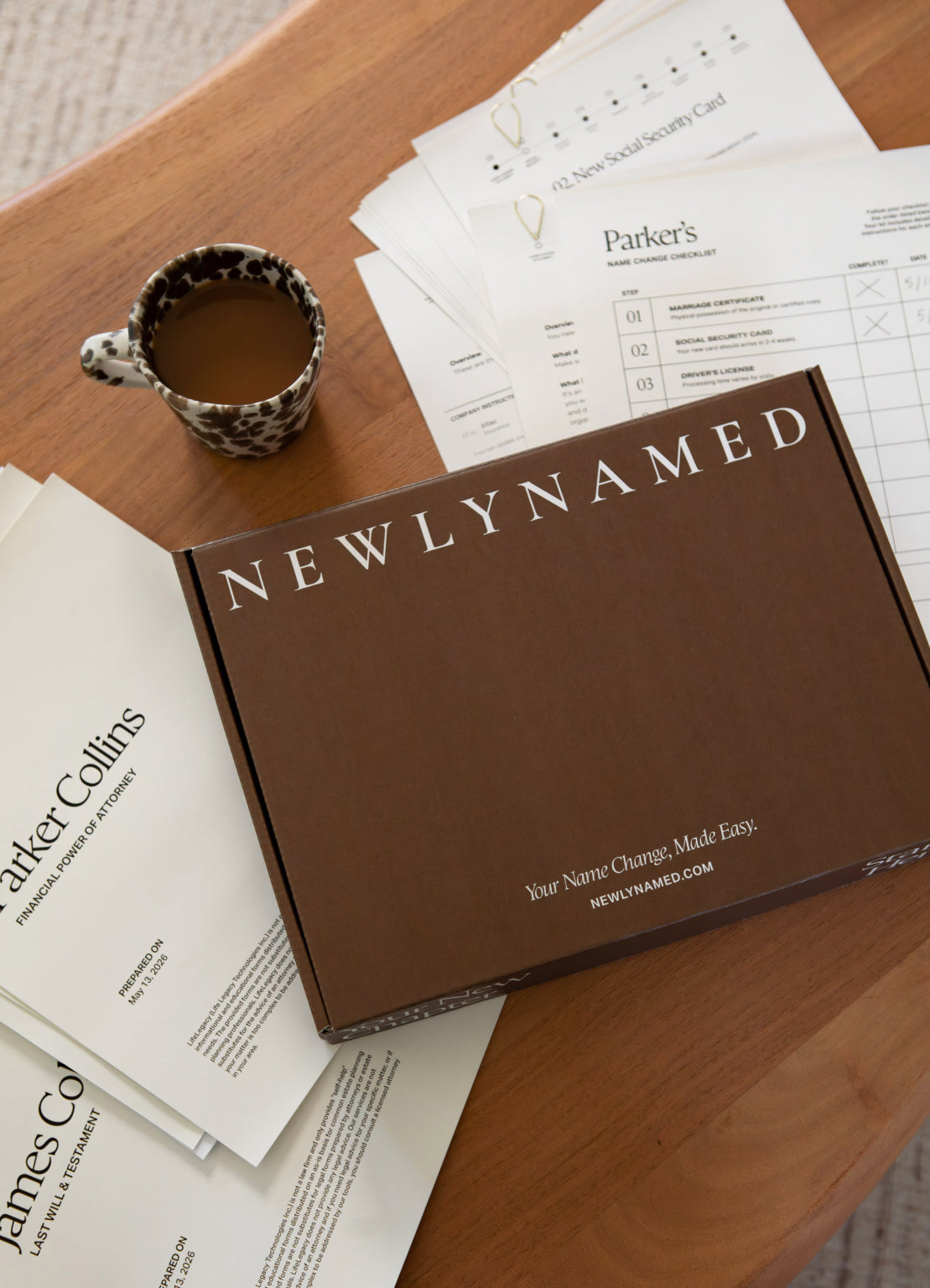 A brown NEWLYNAMED Box name change kit sits on a wooden table surrounded by printed documents and a small patterned cup.