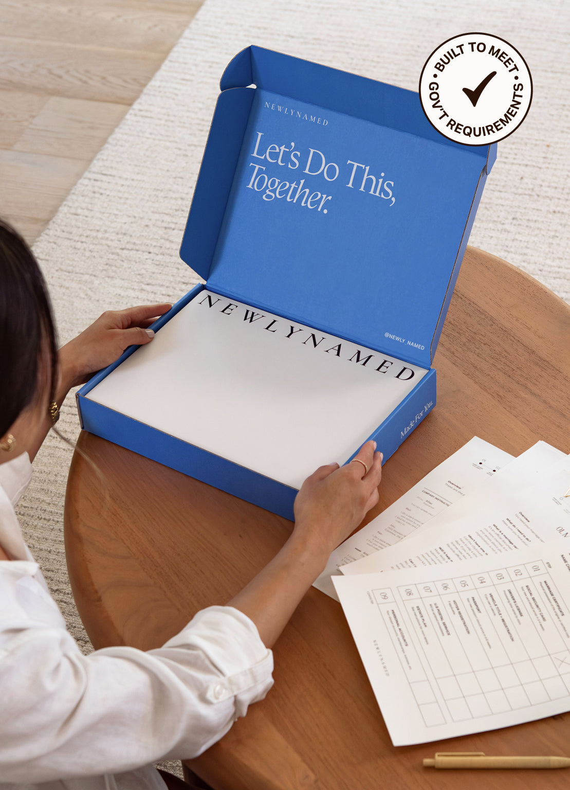 A person seated at a wooden table opens a blue NEWLYNAMED BOX name change kit  containing a personlized name change kit. Papers and a pen are on the table, and a circular badge in the corner reads 'BUILT TO MEET GOVERNMENT REQUIREMENTS.