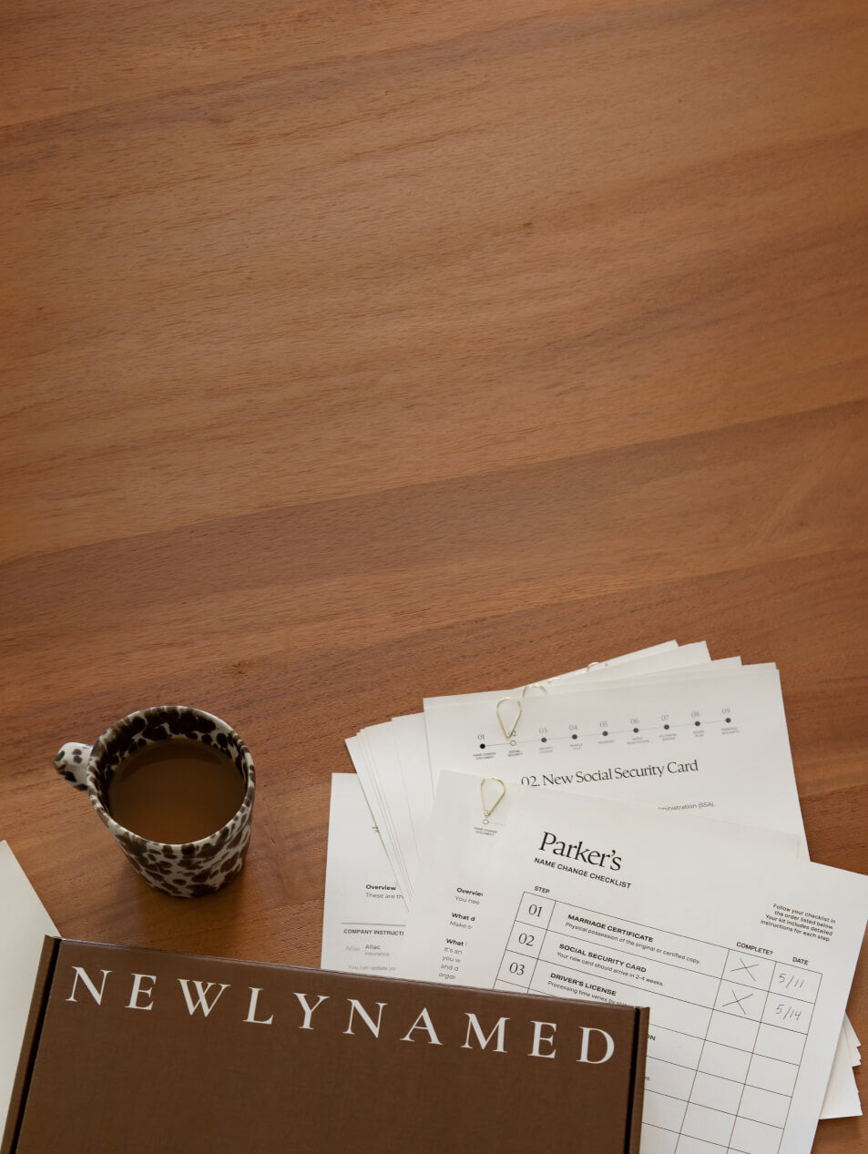A top-down view of a wooden table featuring a dark brown "NEWLYNAMED" box in the bottom left corner next to a black-and-white patterned coffee mug. A stack of paper checklists sits to the right, with the top sheet titled "Parker's Name Change Checklist" showing hand-written checkmarks.