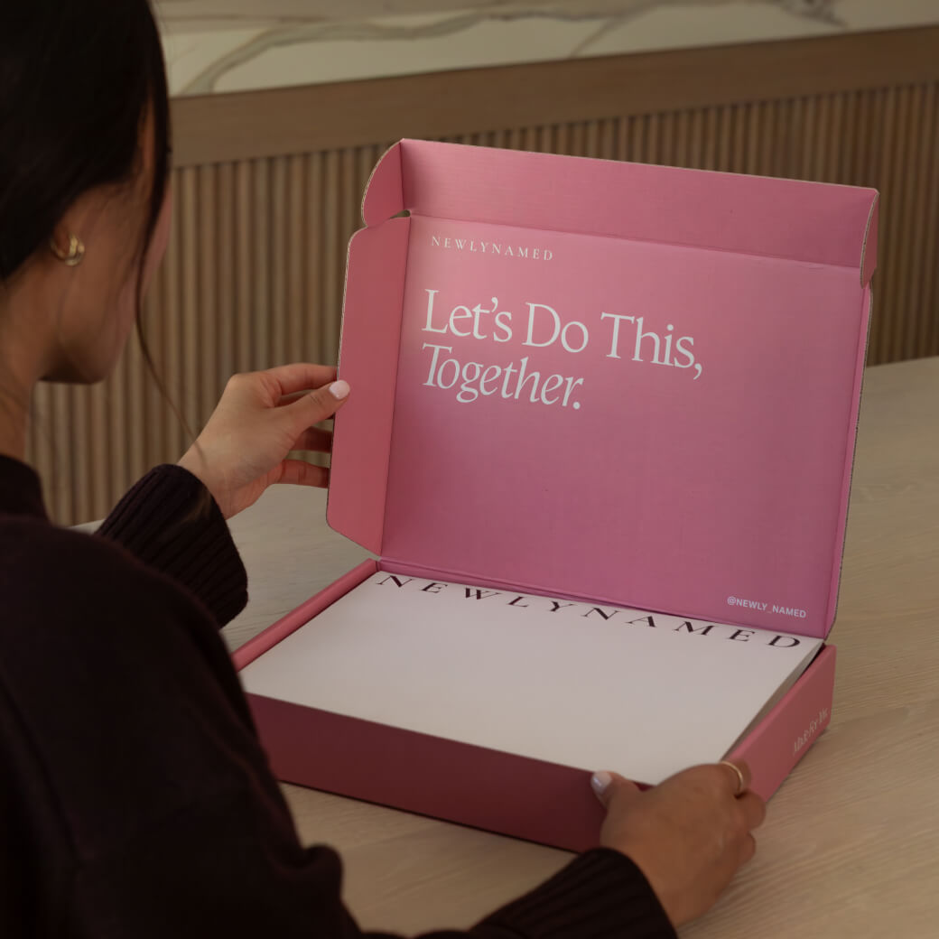 An over-the-shoulder view of a person opening a light pink box. The inside lid of the box displays the words 