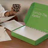 An open bright green box sitting on a wooden table with the white text "Let’s Do This, Together." on the inner lid. In the background, a person holds a printed document next to a black-and-white speckled mug.