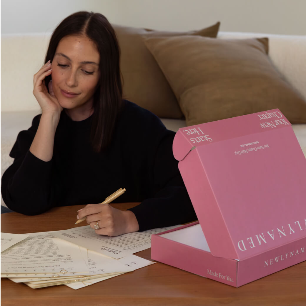 A woman with dark hair sits at a wooden table, resting her chin on her hand while filling out a name change document with a pen. An open pink 