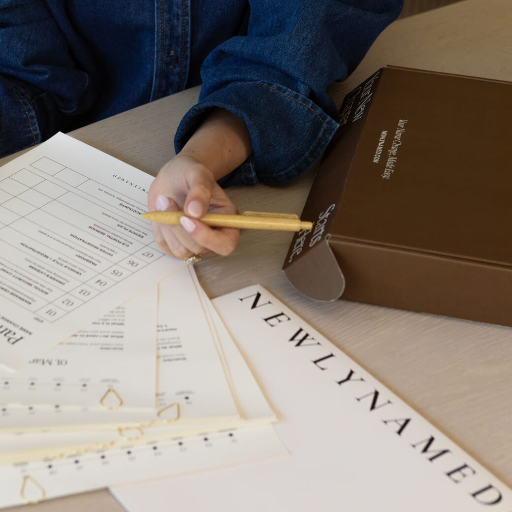 A person wearing a denim shirt holds a tan pen over printed forms on a light-colored wooden table. A closed dark brown 
