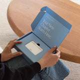 An over-the-shoulder view of a person holding an open blue NewlyNamed gift card box. The inner lid reads "Your New Chapter Starts Here." in white text, showcasing a white "NEWLYNAMED Gift Card" nested inside the custom insert.