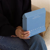 A person sitting and holding a small, partially open blue "NEWLYNAMED" gift box in their hands. The box features clean white text and is designed to hold a gift card.
