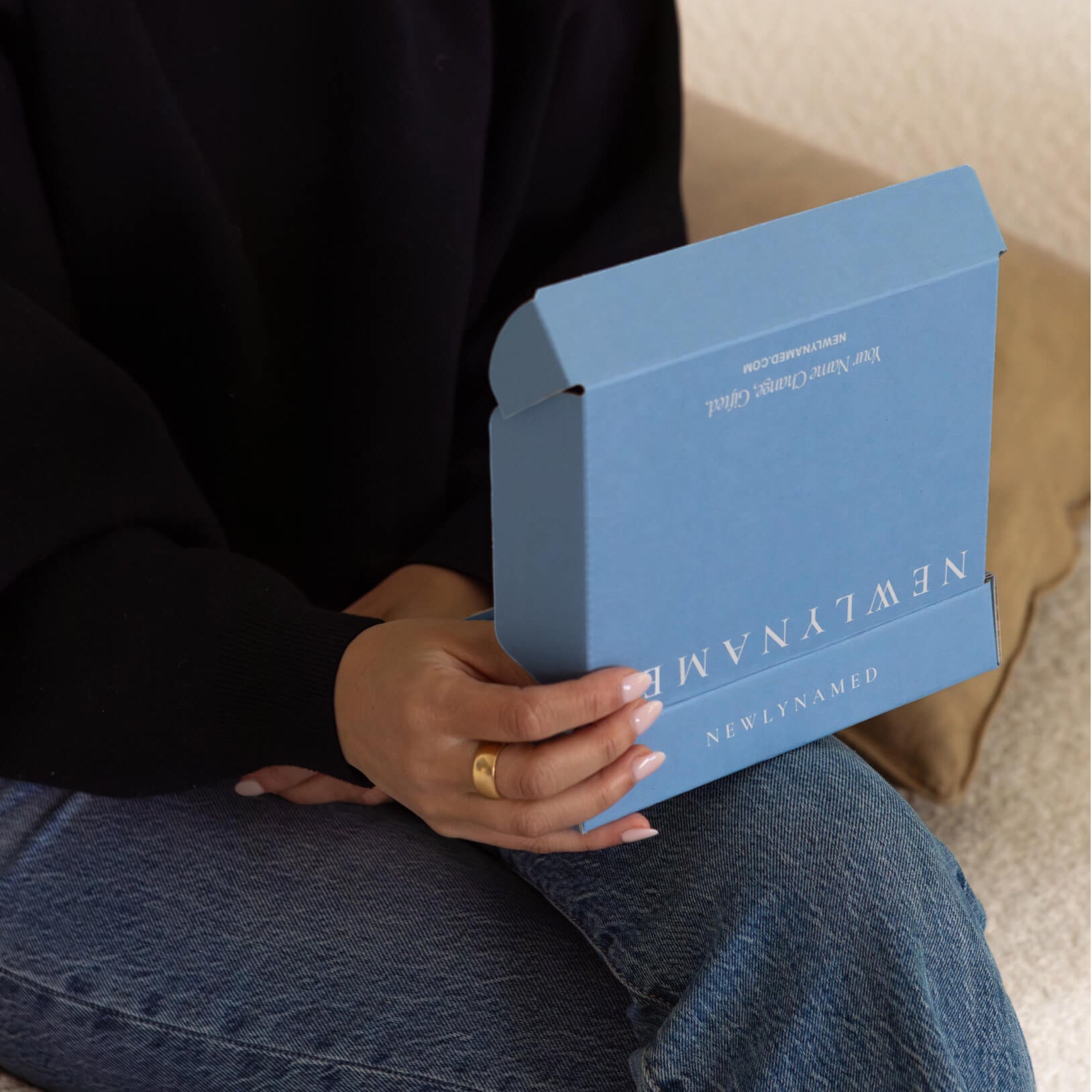 A person sitting and holding a small, partially open blue "NEWLYNAMED" gift box in their hands. The box features clean white text and is designed to hold a gift card.