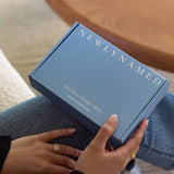 A close-up view of a person holding a closed blue newlynamed gift box on their lap. The top of the box features "NEWLYNAMED" printed in large white serif letters, with "Your Name Change, Gifted." and "NEWLYNAMED.COM" printed along the bottom edge.