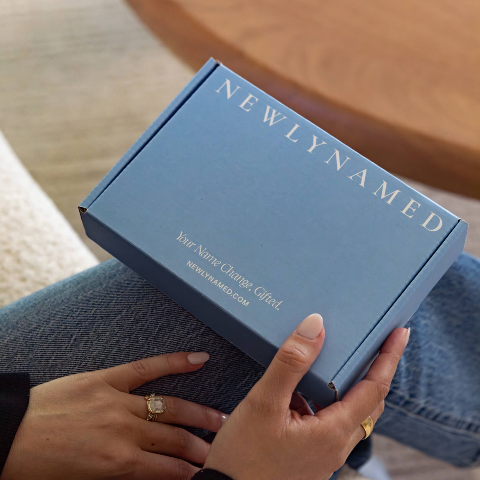 A close-up view of a person holding a closed blue newlynamed gift box on their lap. The top of the box features "NEWLYNAMED" printed in large white serif letters, with "Your Name Change, Gifted." and "NEWLYNAMED.COM" printed along the bottom edge.